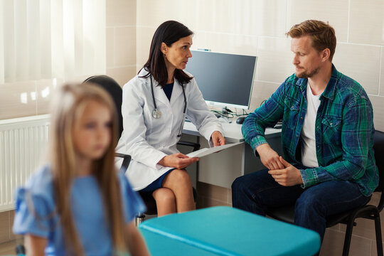 Defocused Little Girl Sitting On Couch In Doctors Office While Female Pediatrician Explaining Diagnosis To Her Frustrated Father In Background