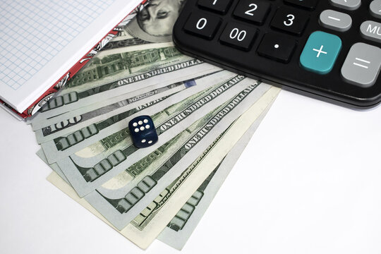 On a light background are dollars on top of which is a blue dice with a calculator and Notepad.
