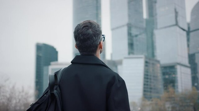 Man in coat looking at the skyscrapers pan shot right to left. Gimbal back shot of businessman in eyeglasses near skyscrapers in business city