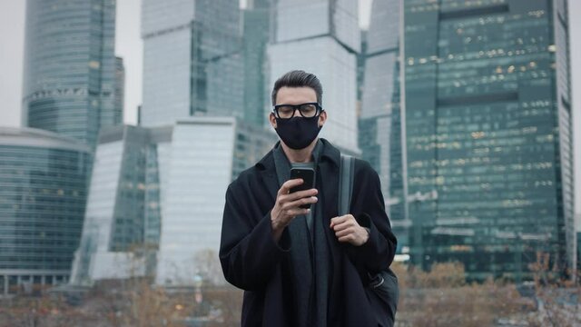 Man in black coat, eyeglasses and black protective mask walking on background of business skyscrapers in Moscow, slow motion. Middle shot of young man in the city business area