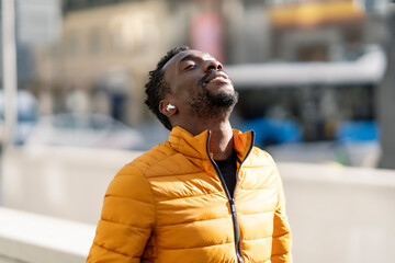 African man listening to music and breathing fresh air outdoors standing in the city - obrazy, fototapety, plakaty