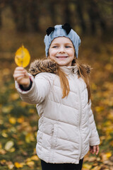 A beautiful cute girl, a child in a white jacket of preschool age, stands and poses with a yellow leaf in nature. Autumn portrait, photography.