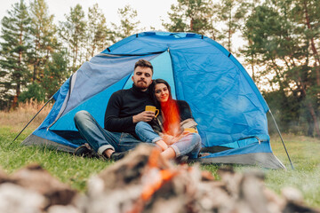 Happiness couple sitting at the entrance of the tent with cups in their hands. Travelers drink tea outdoors