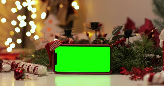iPhone Xs max in Horizontal orientation with green screen on a background of Christmas tree and holiday lights. Santa Claus hat on top. Online store, App promo, ads.
