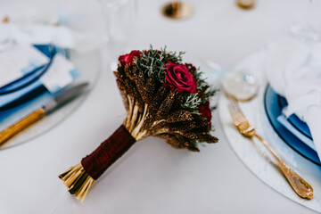 Beautiful and elegant autumn wedding bouquet with dark red roses, pinecones and berry color tape is lying on the decoted table.