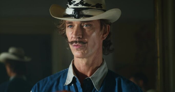 Cinematic close up shot of carefree middle aged happy man with mustache wearing cowboy hat is smiling in camera in living room at home. Concept of non stereotyped people, identity and diversity.