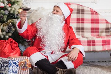 Bearded man dressed in Santa Claus costume sitting on sofa with glass of wine