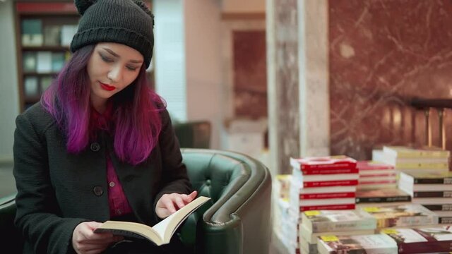 Portrait of stunning asian woman with bright red lips and purple hair reading a book in a bookstore. Concept of hipster style and trends. Millenials lifestyle. teenage student shopping.