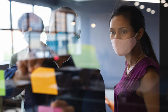 Diverse Business People Wearing Face Masks Writing On Glass Board In Office