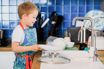 a boy in an apron washes dishes in the kitchen at home.