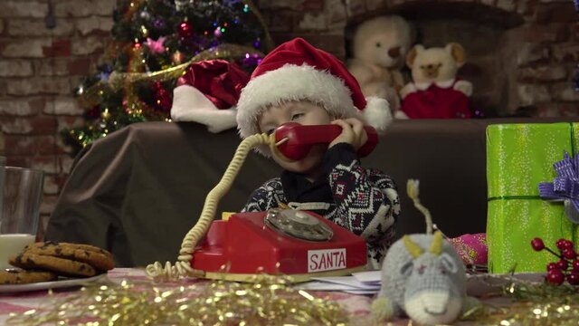 A little boy in a Santa hat sits at the table and calls Santa Claus on an old wired telephone.Santa Claus house.