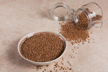 Bowl of dry raw buckwheat groats and glass jar on a light beige background. Cooking buckwheat porridge concept.