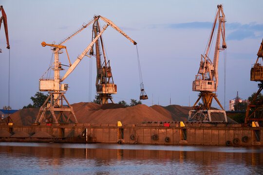 level-luffing bulk-handling cranes load sand onto a barge in a river port in the evening light