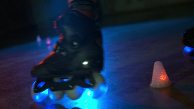 Close-up of teenager trick roller skating in dark studio with colourful lights. Millennial doing stunt show riding around obstacles, wheels flashing blue. Concept of lifestyle, nightlife, extreme.