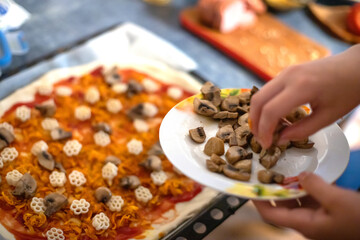 Hand adding mushrooms to the pizza. chef makes pizza heart for Valentine's Day holiday at home on table. Lifestyle concept. Home-cooked food on self-isolation during coronavirus