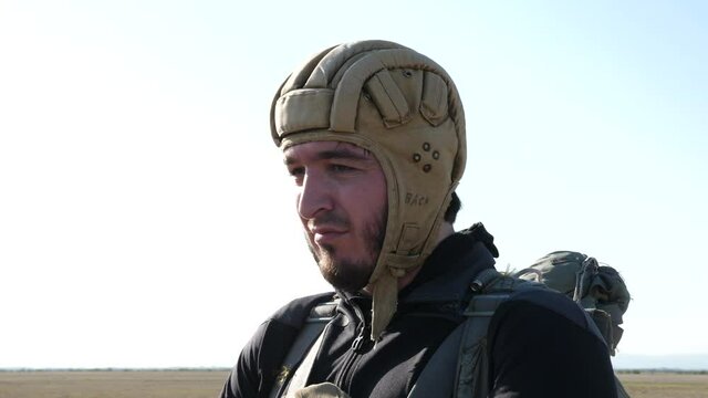 Nervous man with black beard wearing military protective helmet and parachute bag prepares for jump on sunny day closeup
