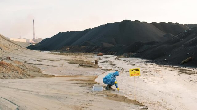 Full shot of ecology expert in protective costume with test tube taking probe for quality control from dirty contaminated water in toxic dangerous zone