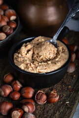 Hazelnut butter in ceramic cup with shelled hazelnuts on wooden background.