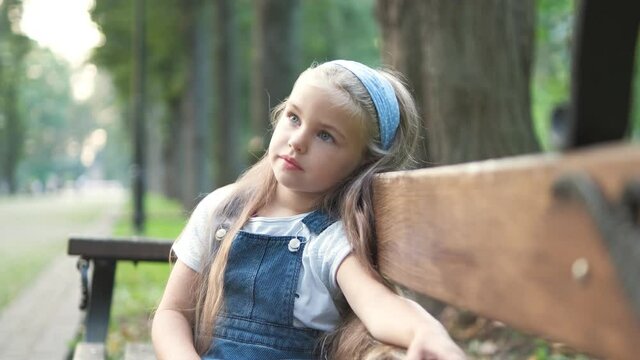 Little child girl sitting alone on a bench in summer park.