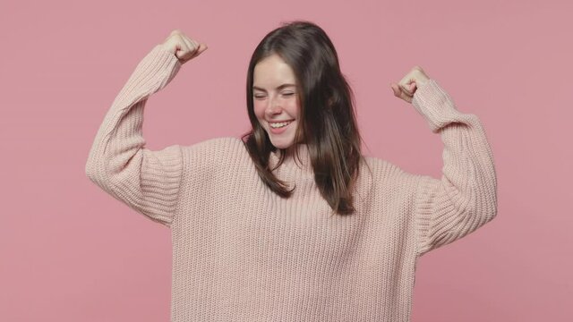 Strong cheerful brunette young woman 20s years old in casual pastel sweater posing isolated on pink color background in studio. People lifestyle concept. Looking camera aside showing biceps mucles