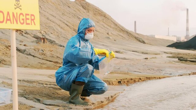 Medium shot of female ecologists in protective suits and respiratory masks collecting material evidence from toxic polluted water into test tubes and placing it in individual packs