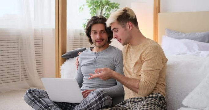 Happy gay couple using laptop sitting in bedroom at home