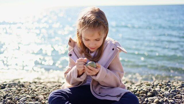 a girl in a coat sits on a pebble sea shore and paints with a stone on a stone.
