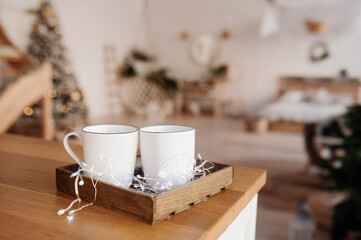 two clear white ceramic mug and garlands on wooden table in christmas decor and scandi style blurred room in pastel colors. selective focus