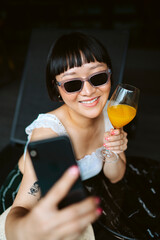 Short hair woman wearing sunglasses taking selfie with orange juice.