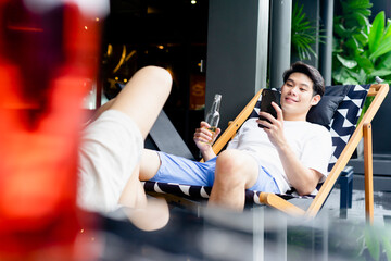 Asian man drinking wine from bottle and using smartphone while laying on beach chair.
