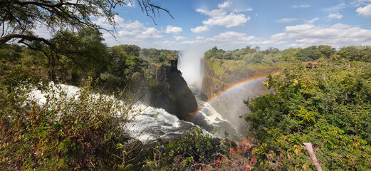 Swirling Zambezi River. Victoria Falls. Rainbow on a background of blue sky with white clouds.