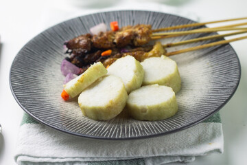 Close up view of Sate Ayam or Indonesian Chicken Satay served pn ceramic grey plate with lontong slices, chilies, shallot, and peanut sauce. Isolate white background.