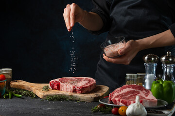 Chef pours sea salt on raw steak on wooden chopped board. Backstage of preparing grilled pork meat...