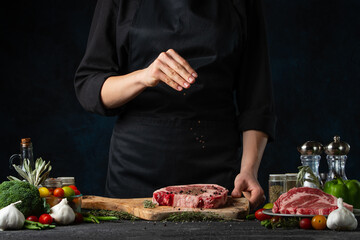 Chef salts raw steak on wooden chopped board. Backstage of preparing grilled pork meat at restaurant kitchen on dark blue background. Frozen motion. Cooking process.