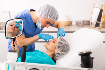 Confident man beautician showing to young female patient results of cosmetology beauty procedure in clinic