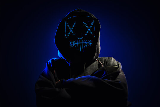 Anonymous Man In Hoodie Hiding Face Behind Neon Glow Scary Mask On Dark Background. Horror Concept