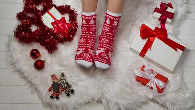 Female legs in warm socks with red white ornaments on a background of fur skin, tinsel and boxes with gifts relaxing at home. Winter holiday Christmas and New Year concept. Close up. Slow motion.