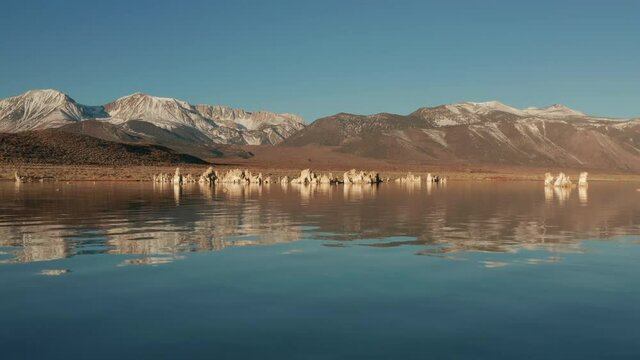 Relaxing nature aerial footage with copy space for background. Drone flying above still lake surface on sunny morning. Snowy mountain peaks and unique tufa formations reflecting in smooth water, 4K