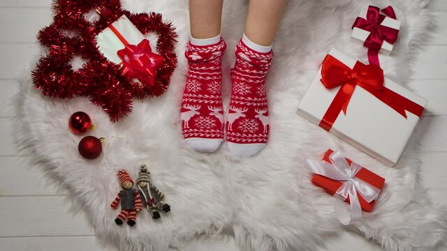 Female legs in warm socks with red white ornaments on a background of fur skin, tinsel and boxes with gifts relaxing at home. Winter holiday Christmas and New Year concept. Close up. Slow motion.