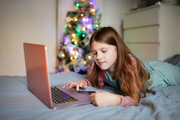Cute girl child with long hair with laptop and credit card makes purchases on the internet, kid online shopping on the bed in the bedroom with a christmas tree