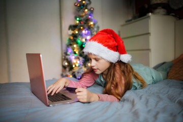 Fototapeta na wymiar Cute little girl child in red santa hat with laptop and credit card makes purchases on the Internet, child online shopping on the bed in the bedroom with a christmas tree