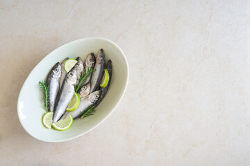 Salted herrings, fresh rosemary, lemon slices on beige bowl over marble background with copy space.