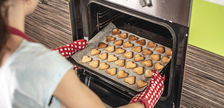 Woman is taking out a baking tray with delicious homemade heart-shaped cookies from the oven. Concept of fresh homemade cakes or a surprise for Valentine's Day
