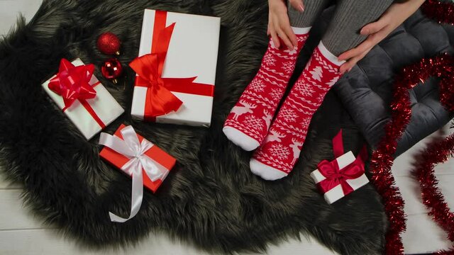 Woman in Christmas warm socks with red and white ornaments sits on a gray fur skin. Presents, jewelry and tinsel are spread around. Holiday date. Merry christmas and new year. Close up. Slow motion.