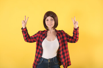 Close-up portrait of a young girl who shows both hands the v-sign symbol and smiles on a yellow background with space for text