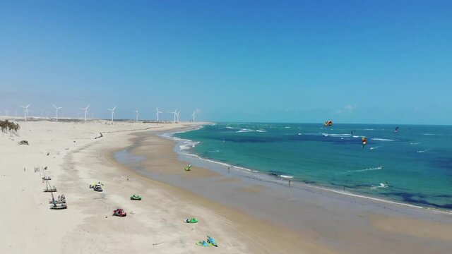 Drone landing on a sandy beach with turquoise ocean, kiteboarding, windmills, rustic fisherman boats and pickup cars in the background. Kitesurfing paradise near Taiba in Brazil.