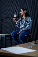 Obraz na płótnie Canvas Happy cheerful pretty smiling of portrait of young Asian woman look at the smartphone vocalist Wearing Headphones recording a song front of microphone in a professional studio