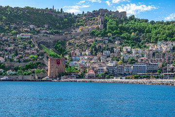 Fototapeta premium View from the sea to the Old City of Alanya (Turkey) with the Red Tower on the background of turquoise water. Panorama of a town with modern buildings on the hillside and an ancient castle at the top