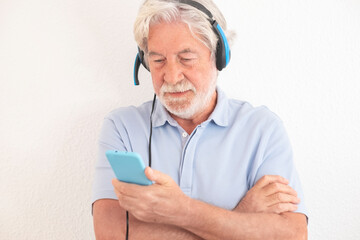 Serious senior white haired  man with headphones standing against white background looking at his smart phone in video call with family. Old people using new technology