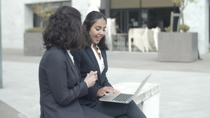 Happy business colleagues sitting outdoors, working with laptop and talking. Professional brunette businesswomen discussing project together. Static camera. Teamwork and digital technology concept - Powered by Adobe